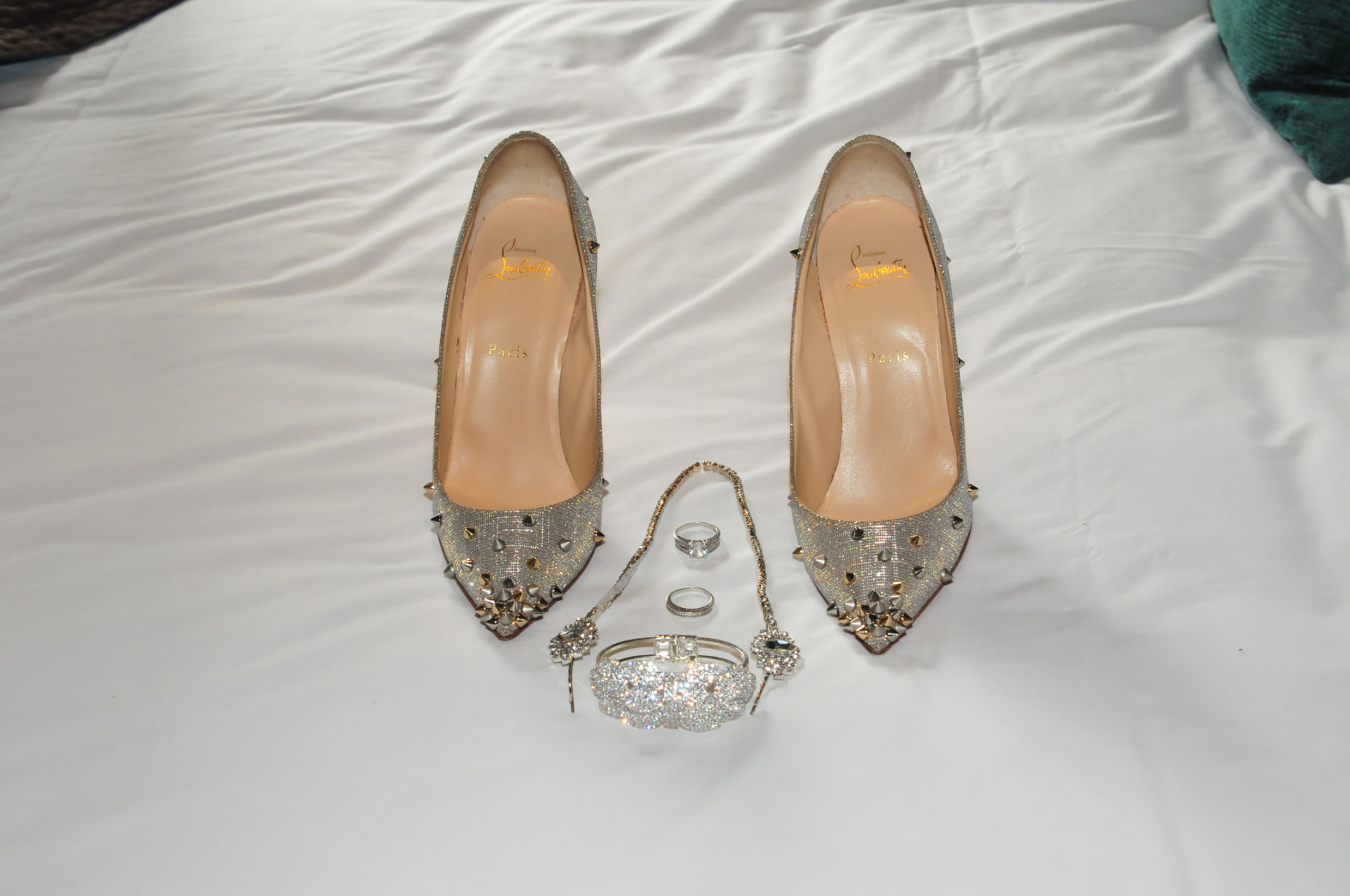 Flatlay of glittered bridal heels, wedding rings, bracelet and necklace on white bedding