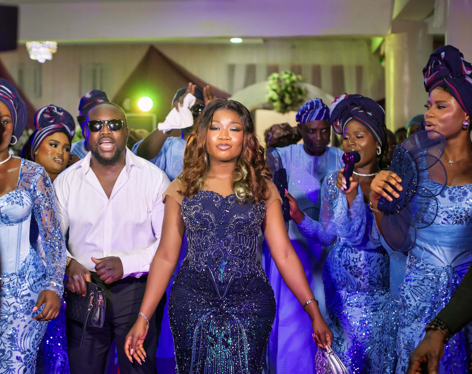 Bride in a navy sequinned gown leading a grand reception entrance with family in blue aso-ebi