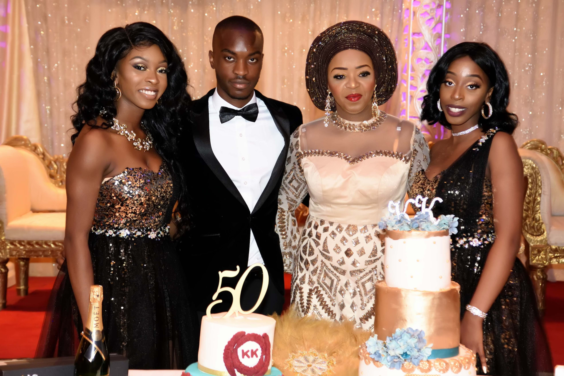 Family in black-tie and gold beaded attire posing beside tiered cakes at a 50th birthday celebration