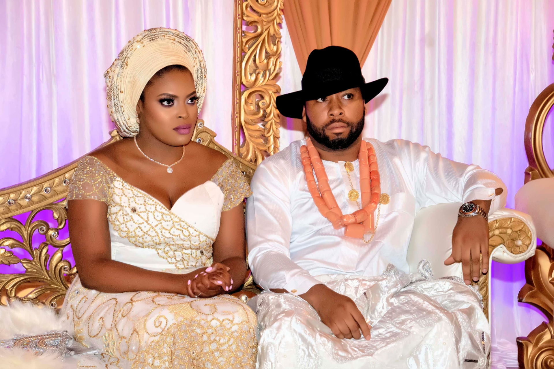Bride in a white and gold beaded gown with gele beside a groom in white agbada and coral beads