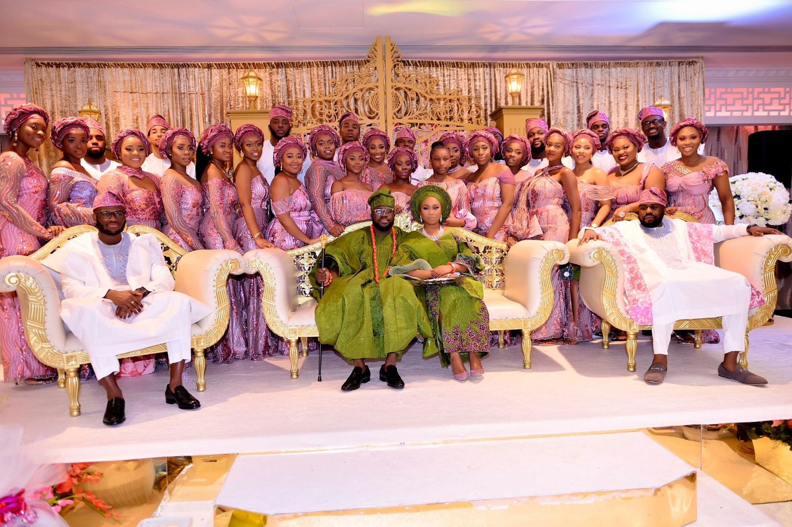 Couple in emerald green agbada and gele seated with a large bridal party in pink aso-ebi on a gold stage