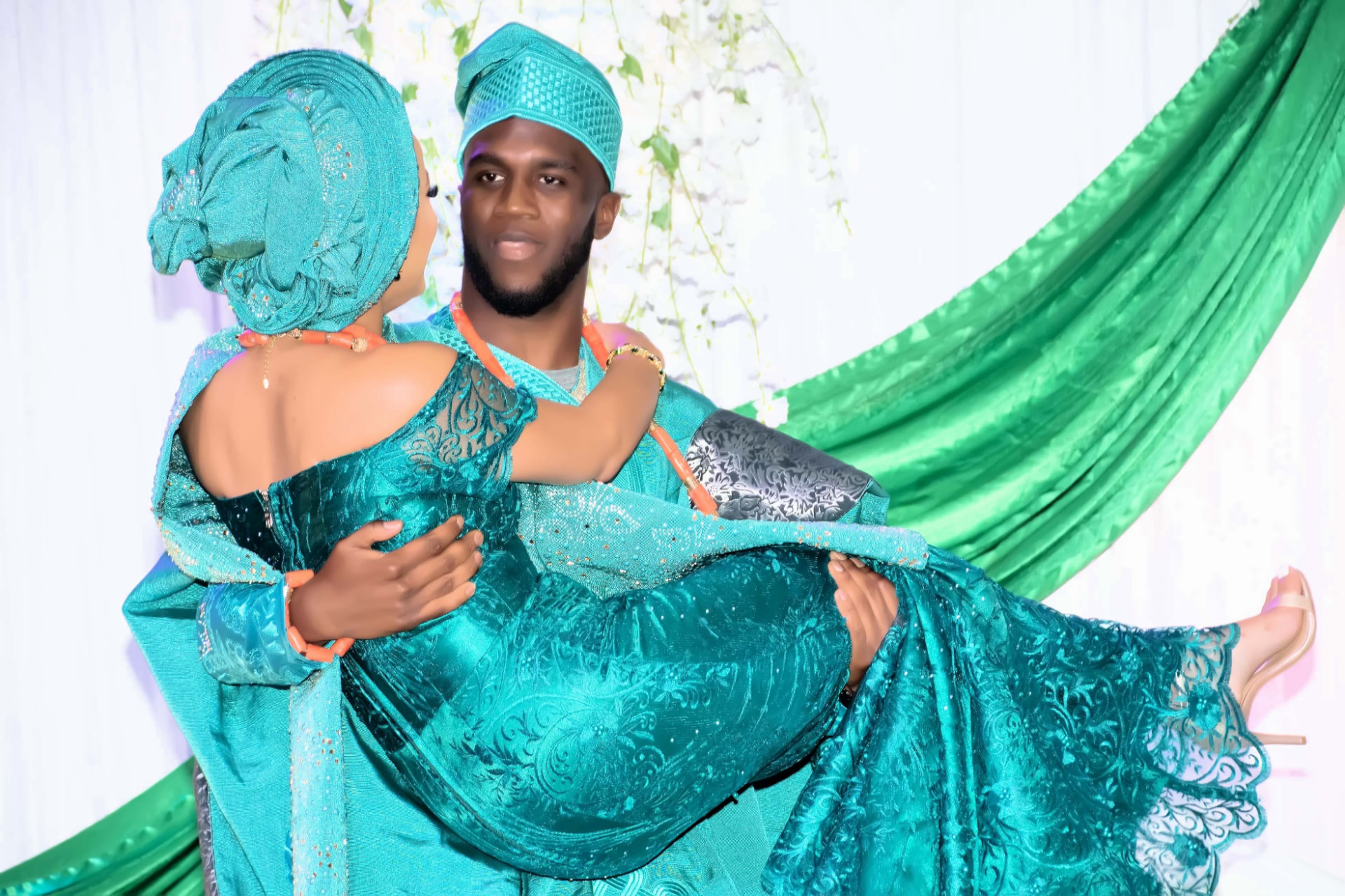 Groom in teal agbada carrying his bride in matching teal lace in front of a green and white backdrop