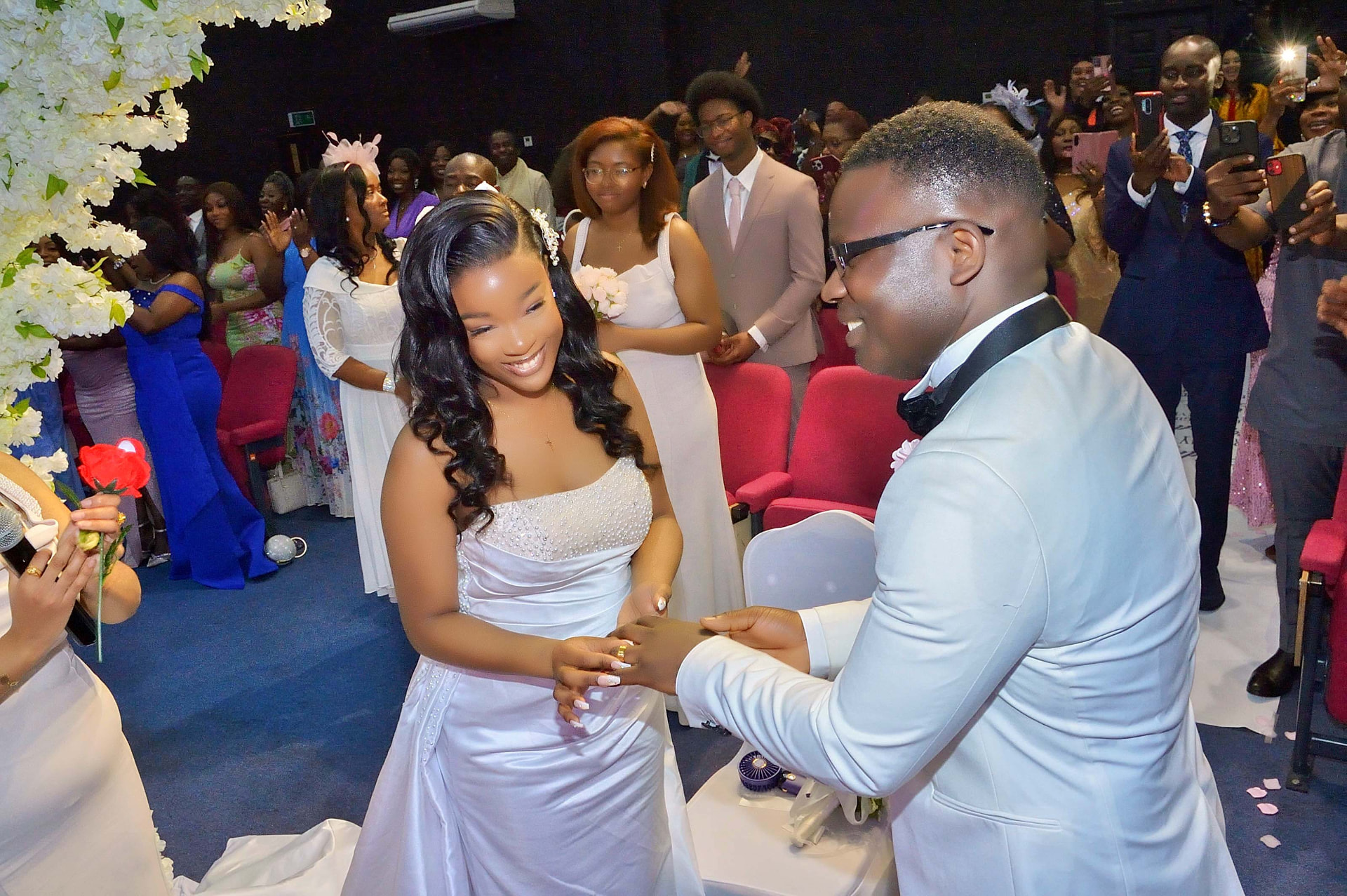 Groom in a white tuxedo placing the ring on his bride's finger as guests applaud during a church wedding